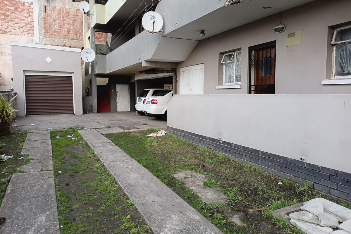 Rear exterior view of an apartment building showing a concrete driveway, garage access, and visible structural wear on the balcony facade.