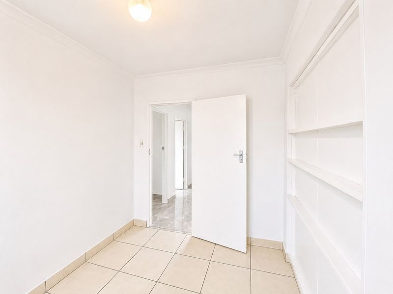 A bright, empty hallway featuring neutral tiled flooring, built-in wall shelving, and coved ceiling details, leading into an adjacent tiled corridor.