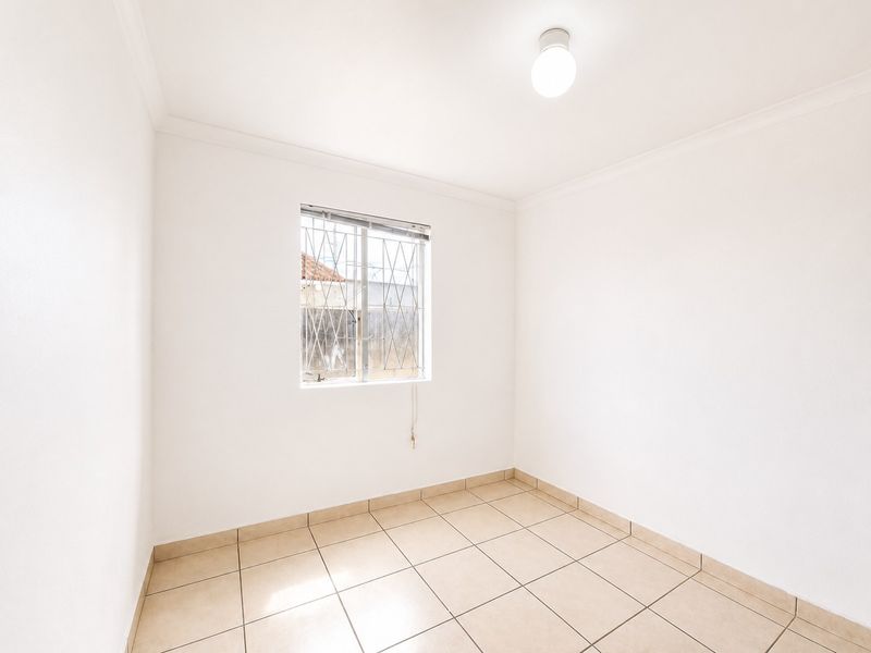 A clean, empty bedroom featuring neutral tiled flooring, white walls, and a window fitted with security bars.