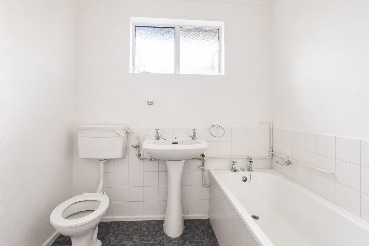 A functional, dated bathroom featuring a pedestal basin, toilet, and bathtub with white wall tiles and exposed plumbing fixtures.