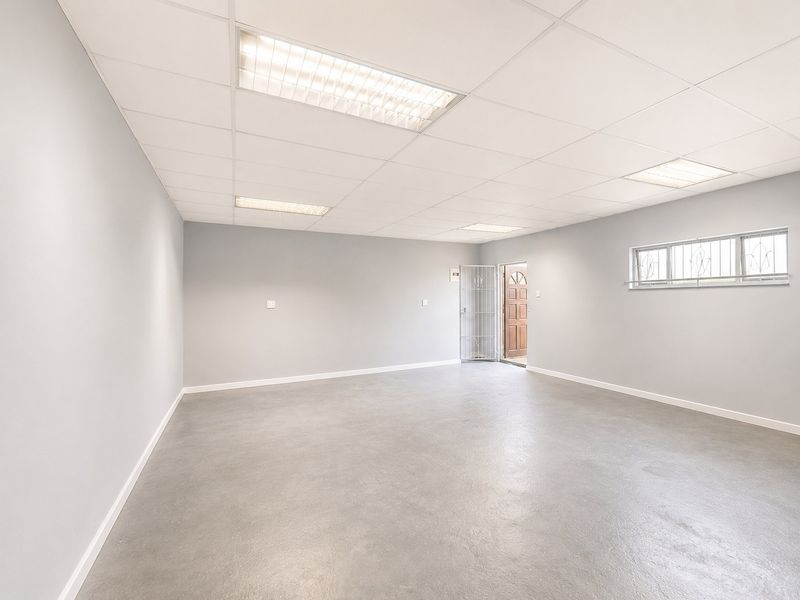 A spacious, empty utility room featuring a suspended ceiling with recessed lighting, concrete flooring, and a secure entrance door with security gate.