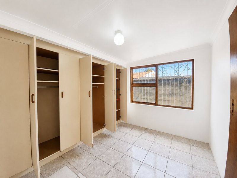 A bright, empty bedroom featuring tiled flooring, built-in cupboards, and a window fitted with security bars.