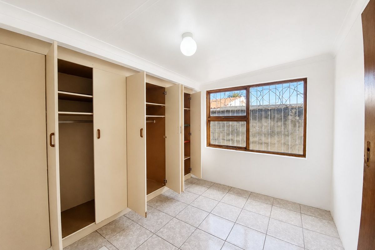 A bright, empty bedroom featuring tiled flooring, built-in cupboards, and a window fitted with security bars.