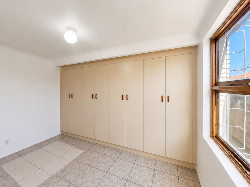 A bright, empty bedroom featuring extensive built-in cupboards, neutral tiled flooring, and a wooden window frame.