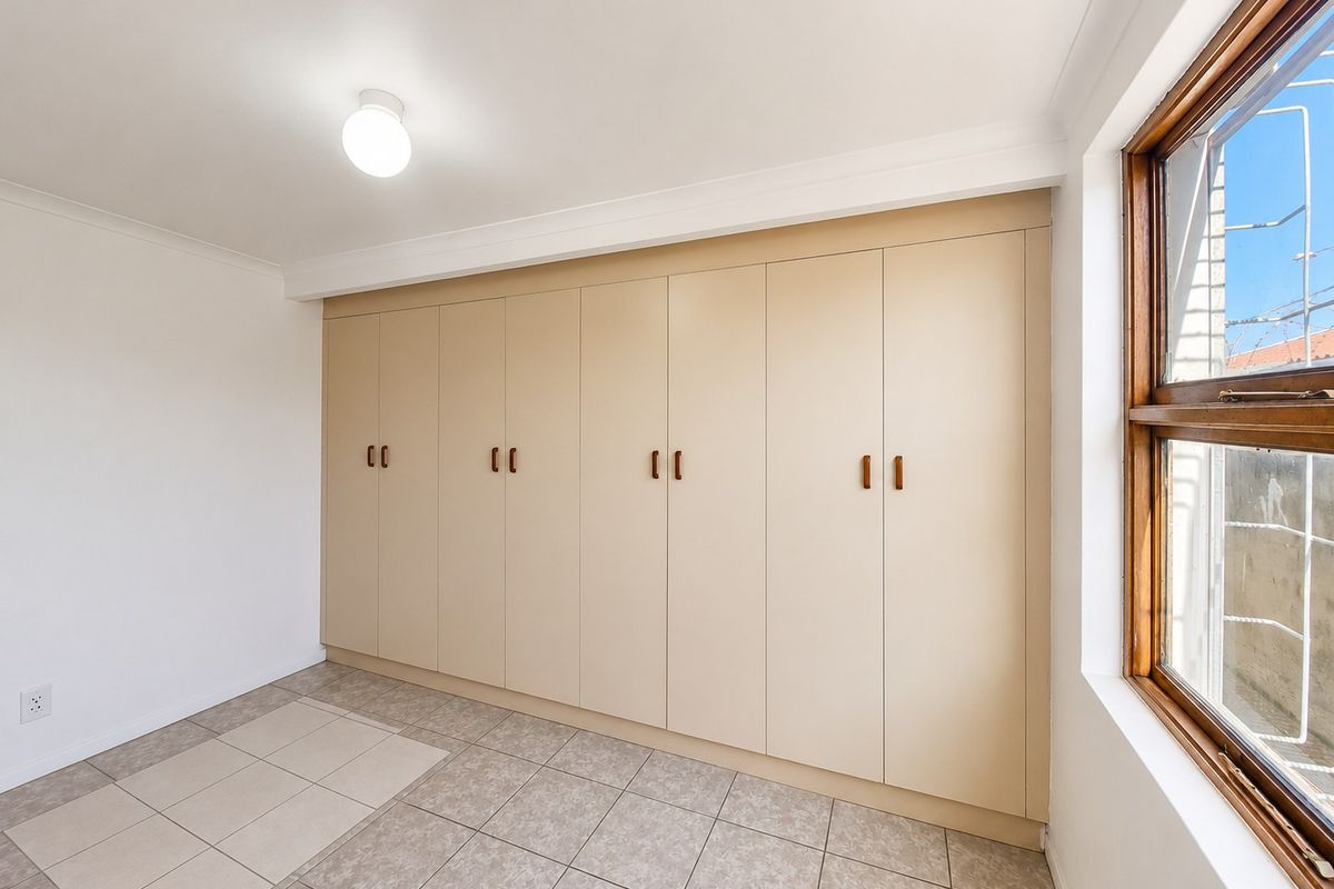 A bright, empty bedroom featuring extensive built-in cupboards, neutral tiled flooring, and a wooden window frame.