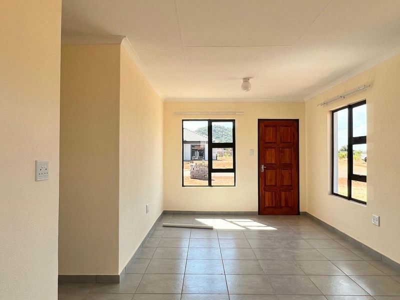 A bright, empty living area featuring neutral walls, tiled flooring, and large aluminium-framed windows providing natural light and access to the exterior.