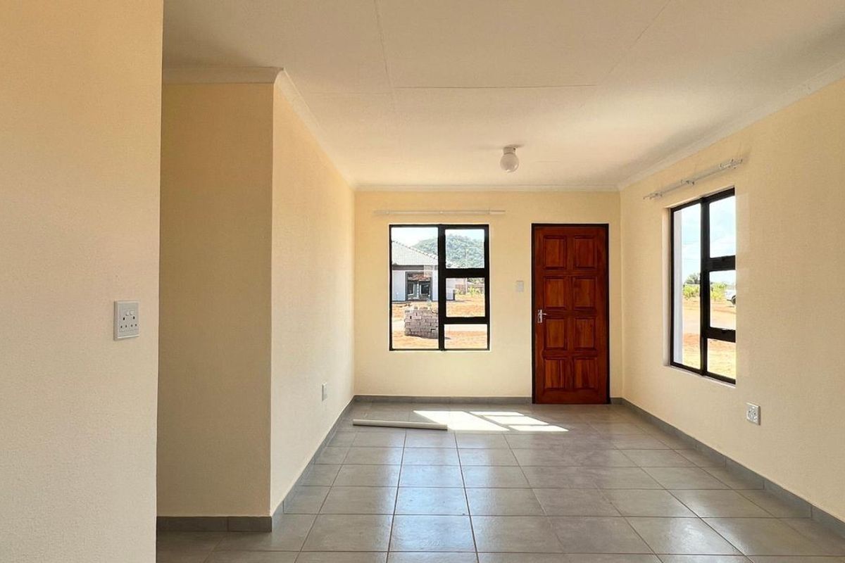 A bright, empty living area featuring neutral walls, tiled flooring, and large aluminium-framed windows providing natural light and access to the exterior.