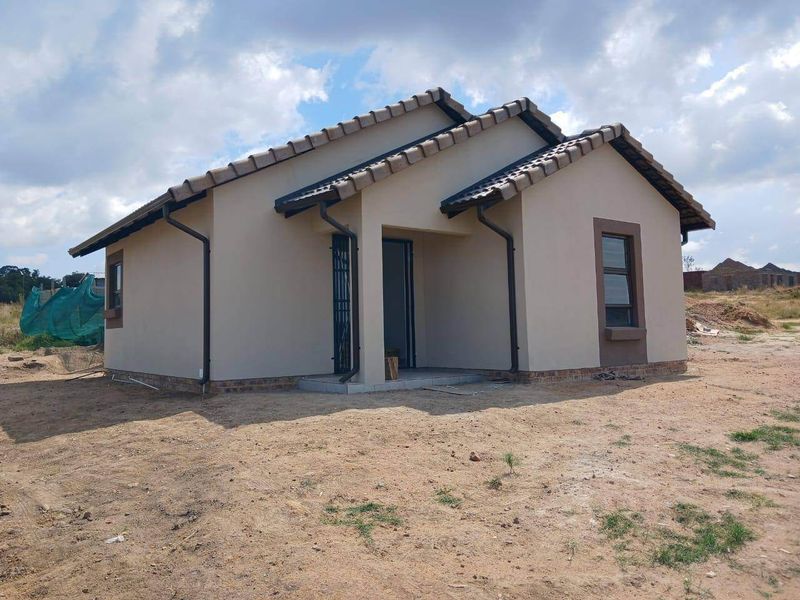 A modern single-story residential house featuring a tiled roof, neutral exterior finish, and security gate at the entrance, set on an undeveloped plot.