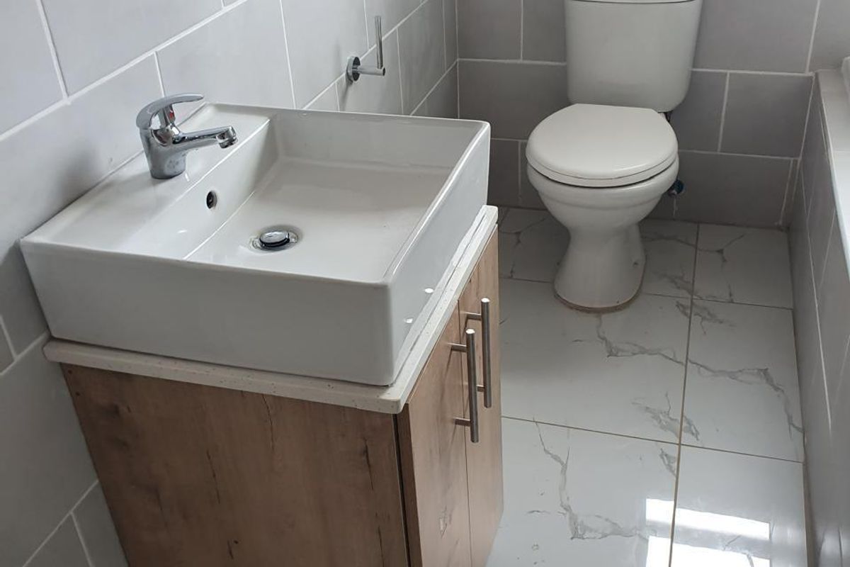 A modern bathroom featuring a wood-finish vanity cabinet with a white ceramic basin, chrome mixer tap, and a standard toilet with marble-look floor tiles.