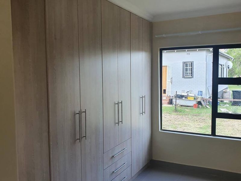 A clean, modern bedroom featuring light wood-finish built-in cupboards, neutral tiled flooring, and a large window providing natural light.