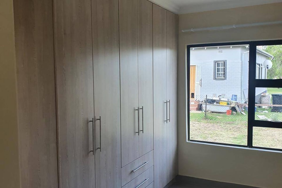 A clean, modern bedroom featuring light wood-finish built-in cupboards, neutral tiled flooring, and a large window providing natural light.