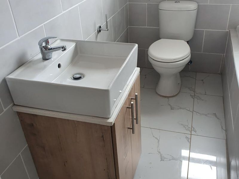 A modern bathroom featuring a wood-finish vanity cabinet with a white ceramic basin, chrome mixer tap, and a standard toilet with marble-look floor tiles.