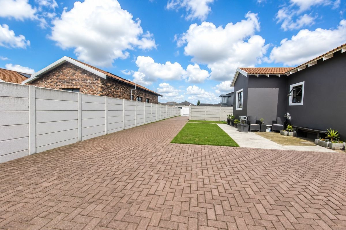 A spacious rear exterior area featuring a large paved driveway, a neat lawn, and a concrete patio adjacent to the main dwelling.