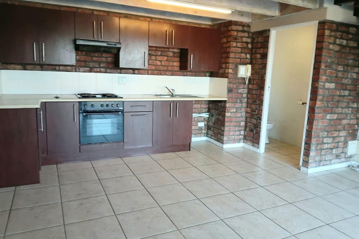 Kitchen featuring dark wood cabinetry, built-in oven and hob, exposed brick walls, and tiled flooring, with an adjacent doorway leading to a bathroom.