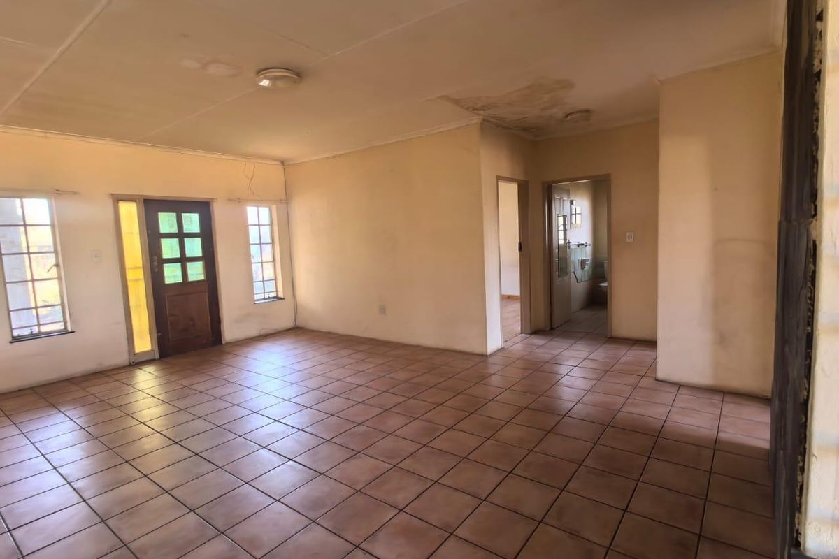 An empty, open-plan living area featuring tiled flooring, natural light through casement windows, and visible ceiling water damage requiring maintenance.
