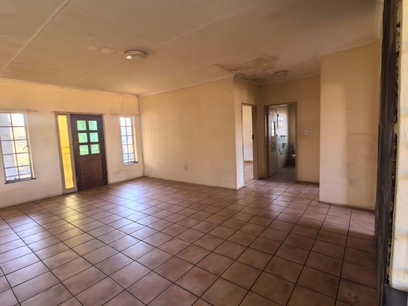 An empty, open-plan living area featuring tiled flooring, natural light through casement windows, and visible ceiling water damage requiring maintenance.