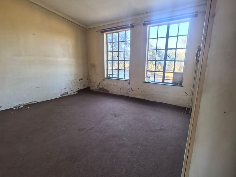 An empty bedroom featuring steel-framed windows and carpet flooring, currently requiring significant wall repairs and cosmetic updates.