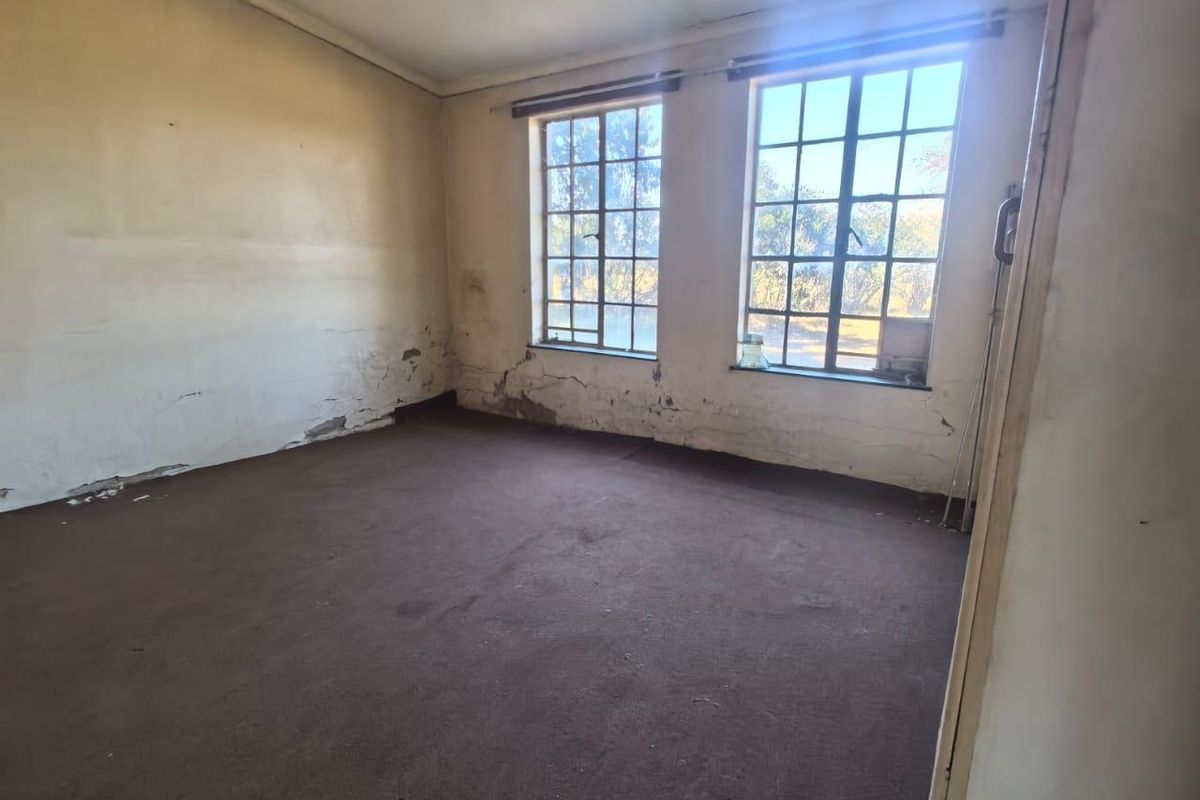 An empty bedroom featuring steel-framed windows and carpet flooring, currently requiring significant wall repairs and cosmetic updates.