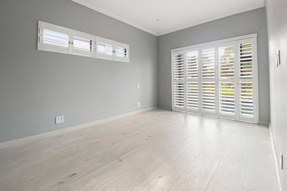 A bright, empty bedroom featuring light-toned laminate flooring, neutral grey walls, and white plantation shutters on both the windows and sliding doors.