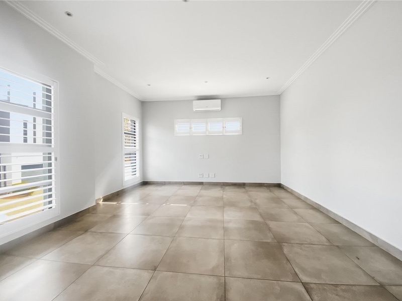 A bright, empty living room featuring large format floor tiles, plantation shutters, and a wall-mounted air conditioning unit.