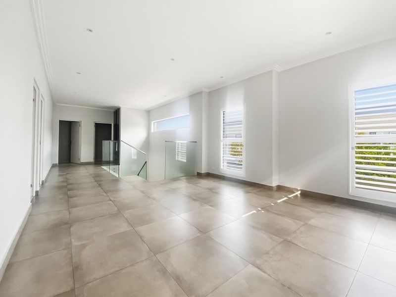 A spacious, modern hallway featuring large format floor tiles, a glass balustrade staircase, and bright natural light through shuttered windows.