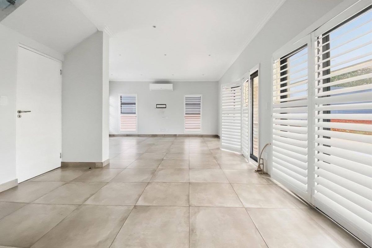 A bright, spacious, and empty living area featuring large format floor tiles, plantation shutters, and a wall-mounted split system air conditioner.
