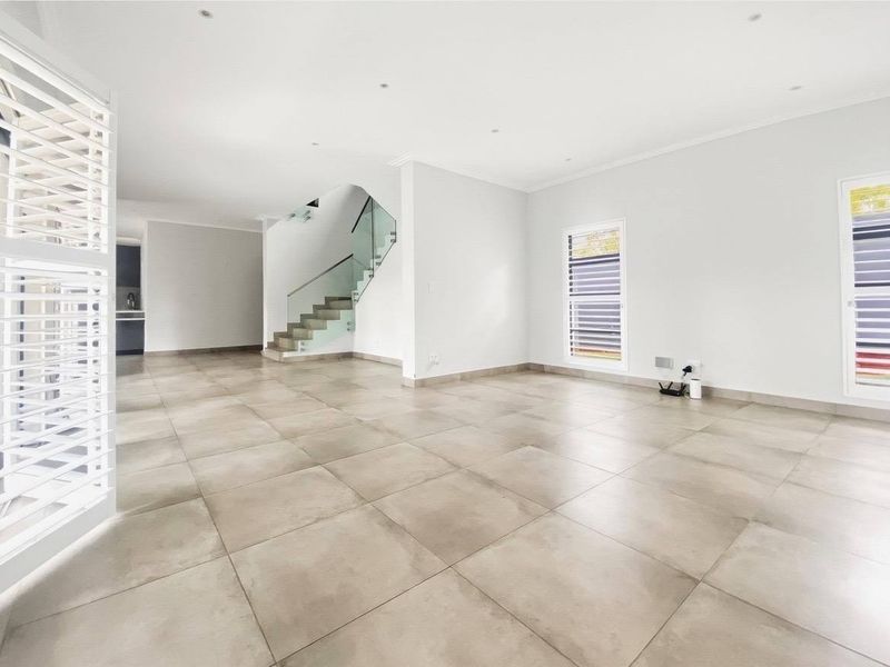 Spacious open-plan living area featuring large format floor tiles, a modern staircase with glass balustrades, and shutter blinds throughout.