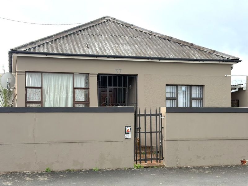 Front elevation of a single-story house featuring a corrugated roof, perimeter wall with security gate, and security bars on windows.