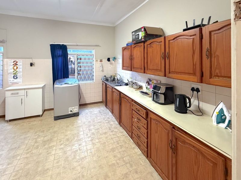 A functional scullery or laundry area featuring wooden cabinetry, laminate countertops, a stainless steel sink, and tiled splashbacks with vinyl flooring.