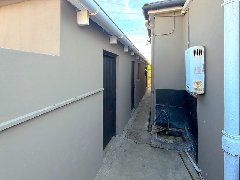 A narrow side passage featuring a concrete walkway, external plumbing, and a wall-mounted gas geyser unit.