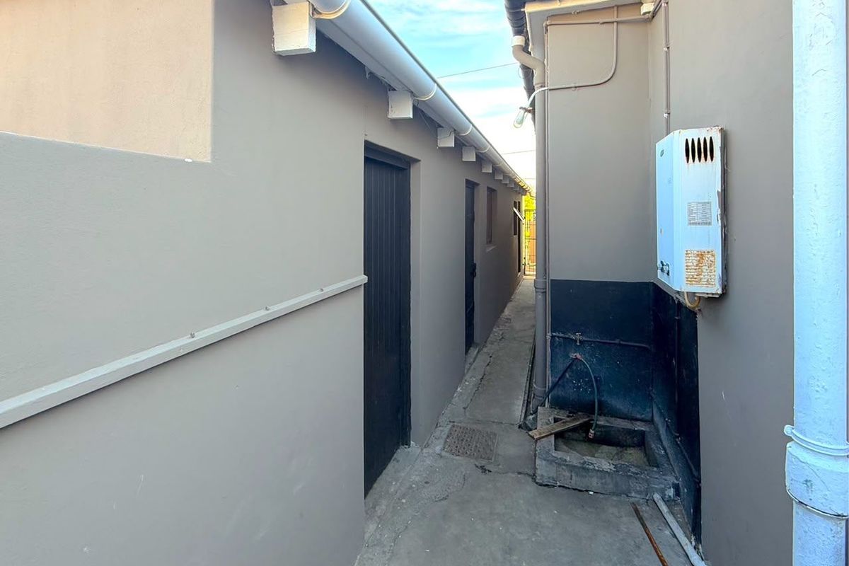 A narrow side passage featuring a concrete walkway, external plumbing, and a wall-mounted gas geyser unit.