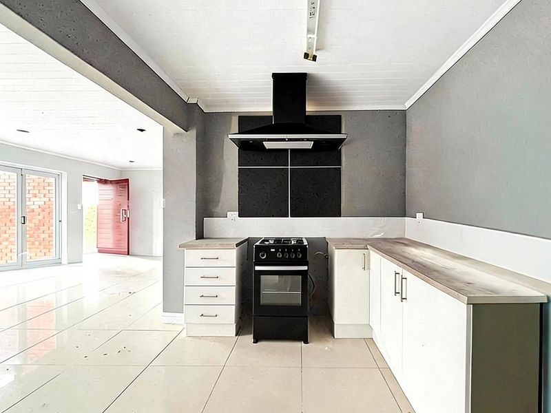 A bright, open-plan kitchen featuring white cabinetry, laminate countertops, a black freestanding stove with extractor fan, and neutral tiled flooring.