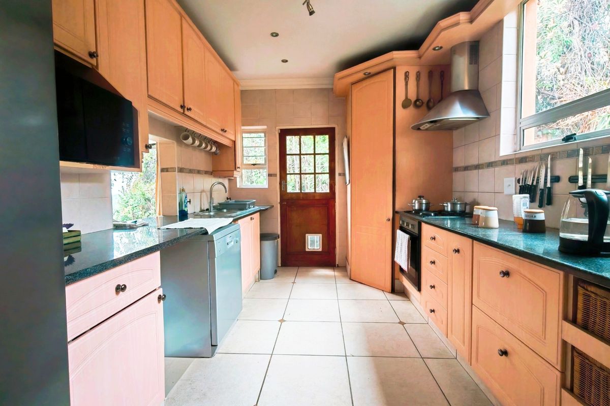 A functional kitchen featuring light wood cabinetry, stone countertops, tiled flooring, and integrated appliances, with direct access to the exterior via a timber door.