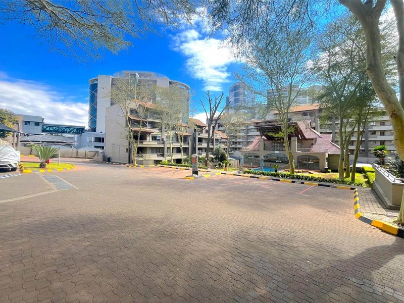 A wide-angle view of the apartment complex exterior, featuring a paved driveway, landscaped grounds, and communal swimming pool area.
