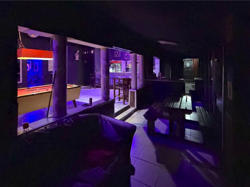 A covered outdoor patio area featuring tiled flooring, support columns, and a built-in bar counter, illuminated with ambient purple lighting.