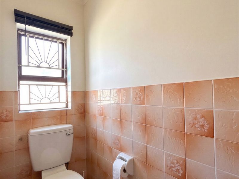 A functional bathroom featuring ceramic wall and floor tiles, a standard toilet, and a window with security burglar bars.