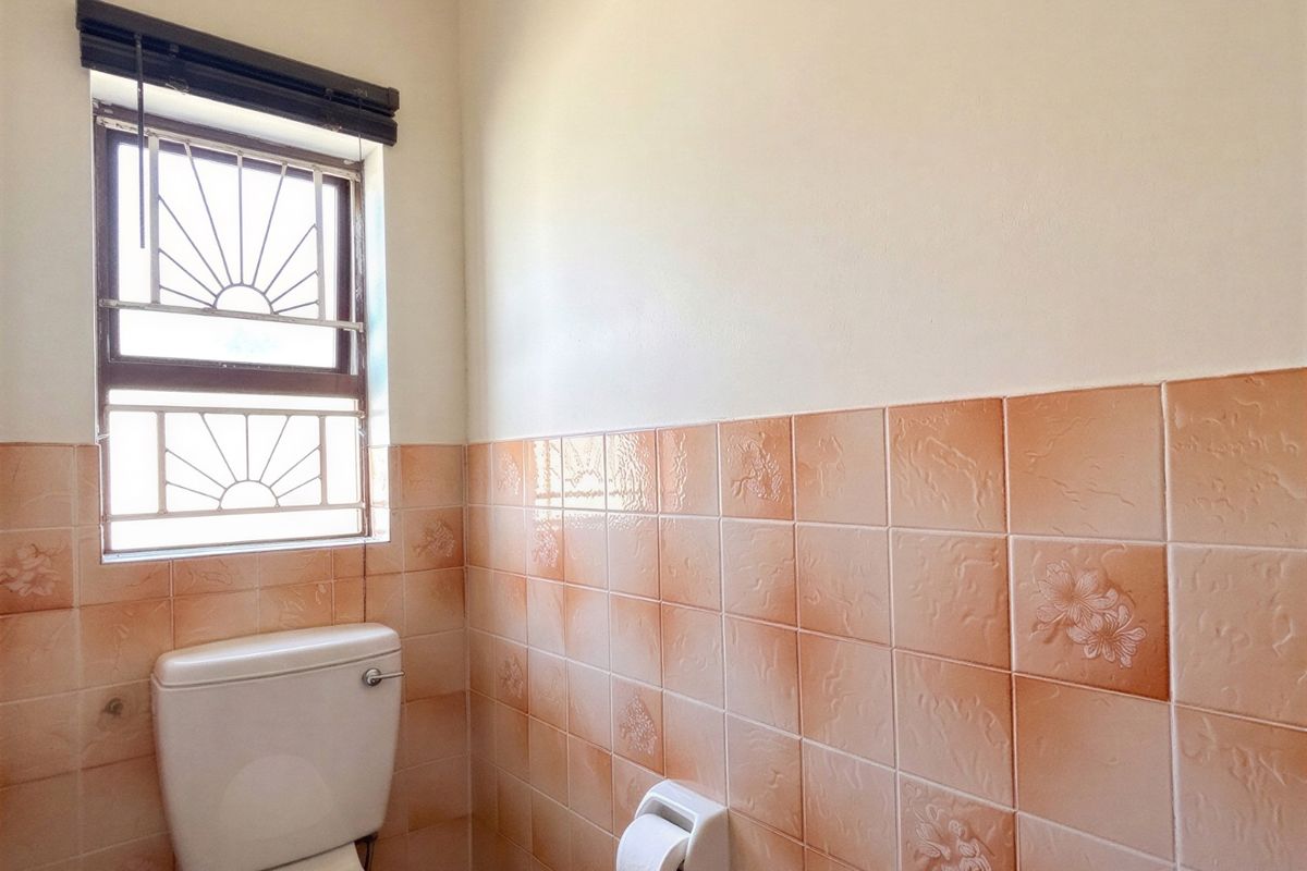 A functional bathroom featuring ceramic wall and floor tiles, a standard toilet, and a window with security burglar bars.