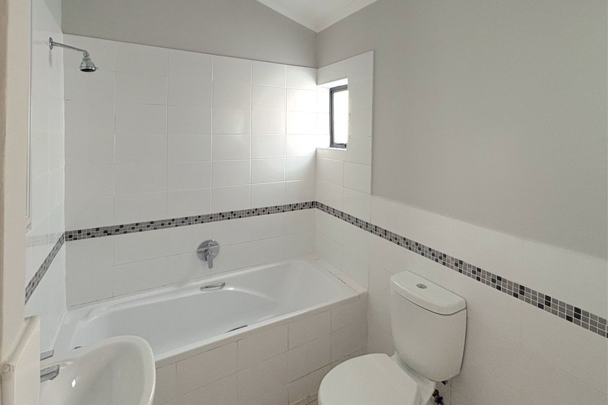A clean, functional bathroom featuring a built-in bathtub with shower, toilet, and washbasin, finished with neutral wall tiles and a decorative mosaic border.