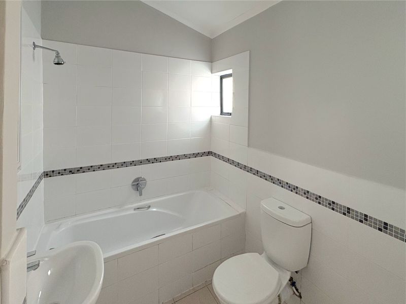 A clean, functional bathroom featuring a built-in bathtub with shower, toilet, and washbasin, finished with neutral wall tiles and a decorative mosaic border.