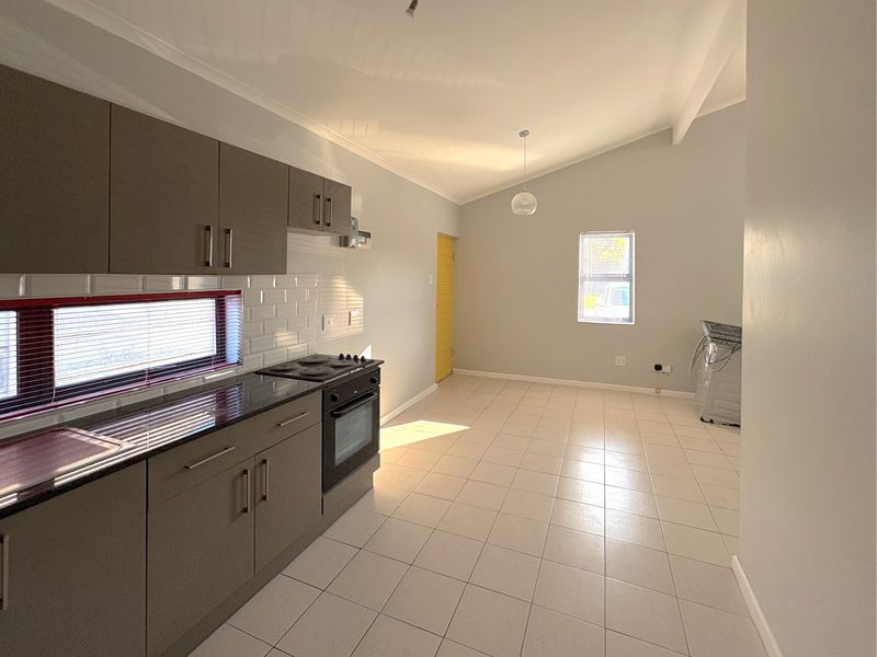 A bright, open-plan kitchen featuring modern grey cabinetry, subway tile backsplash, built-in oven and hob, and neutral tiled flooring under a vaulted ceiling.