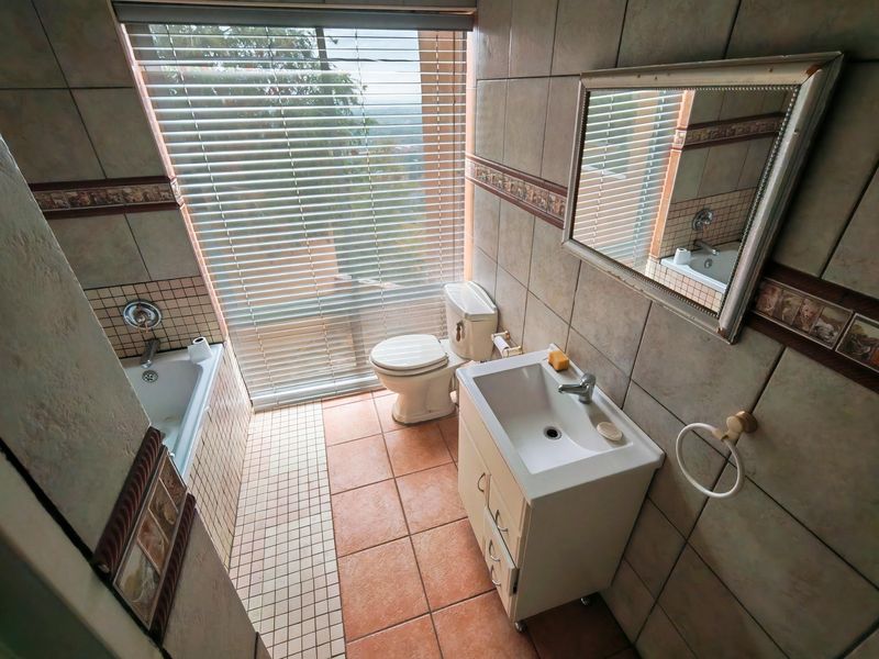 A functional bathroom featuring a built-in bathtub, vanity with washbasin, toilet, and large window with blinds, finished with ceramic wall and floor tiles.