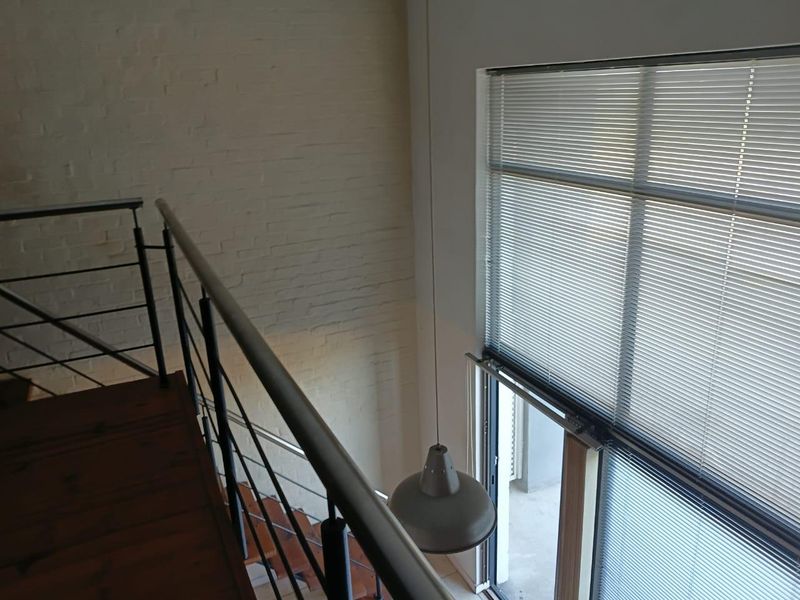 A mezzanine hallway featuring a timber staircase with a modern metal railing, exposed brick walls, and a large window with venetian blinds.