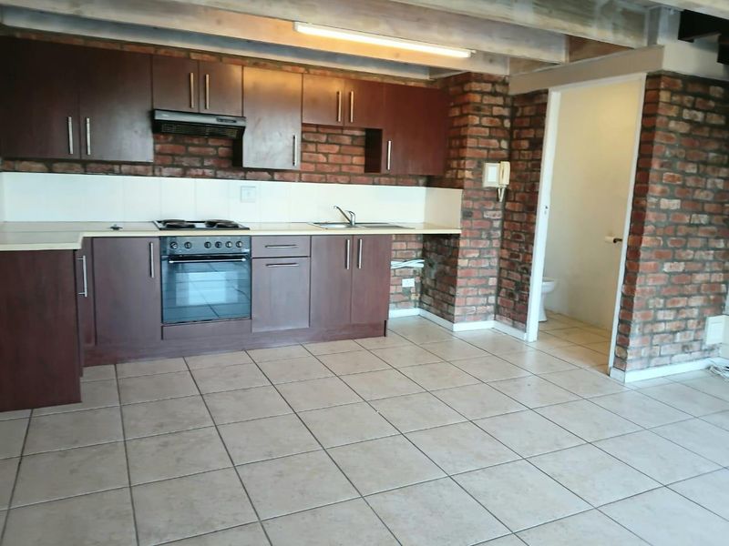 Kitchen featuring dark wood cabinetry, built-in oven and hob, exposed brick walls, and tiled flooring, with an adjacent doorway leading to a bathroom.