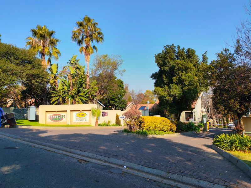 The entrance to Crawford Manor townhouse complex, featuring a paved driveway, security gatehouse, and well-maintained landscaped gardens.