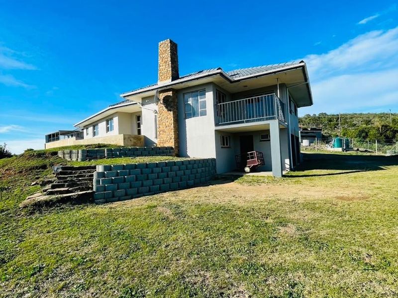 Rear view of a farm-style residence featuring a prominent stone chimney, a balcony, and tiered retaining walls set within a large grassy landscape.