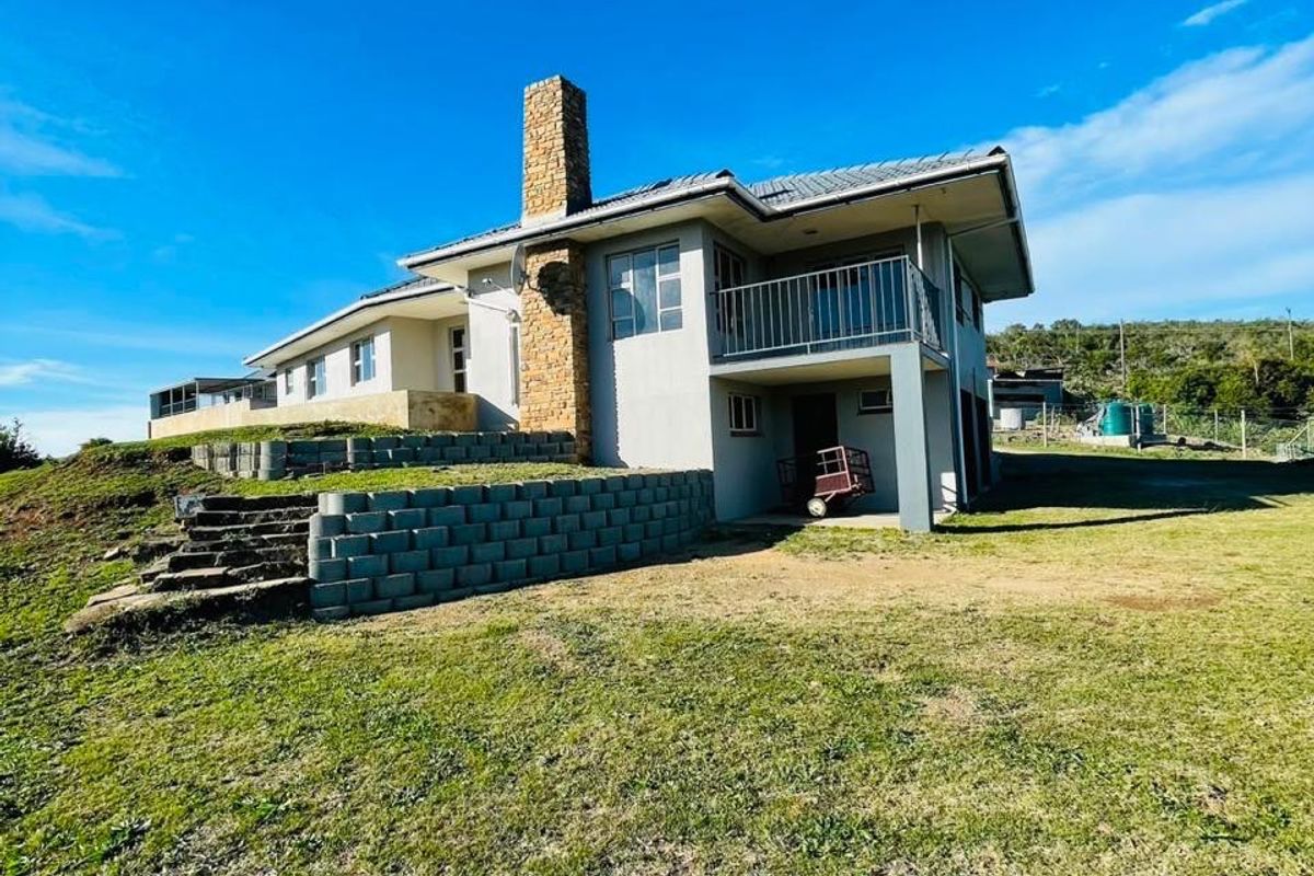 Rear view of a farm-style residence featuring a prominent stone chimney, a balcony, and tiered retaining walls set within a large grassy landscape.