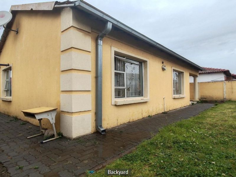 Rear exterior view of the house featuring a paved walkway, downpipe, and boundary wall, showing signs of wear and maintenance requirements.