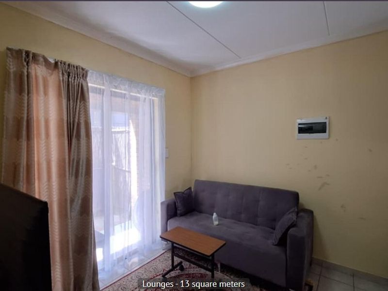 A compact living room featuring tiled flooring, a sliding glass door providing natural light, and a wall-mounted electrical distribution board.
