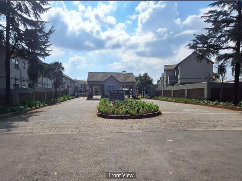 The entrance to Silver Pines complex featuring a paved driveway, security gatehouse, and well-maintained perimeter walls with landscaped garden beds.