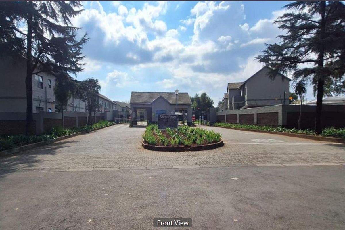 The entrance to Silver Pines complex featuring a paved driveway, security gatehouse, and well-maintained perimeter walls with landscaped garden beds.